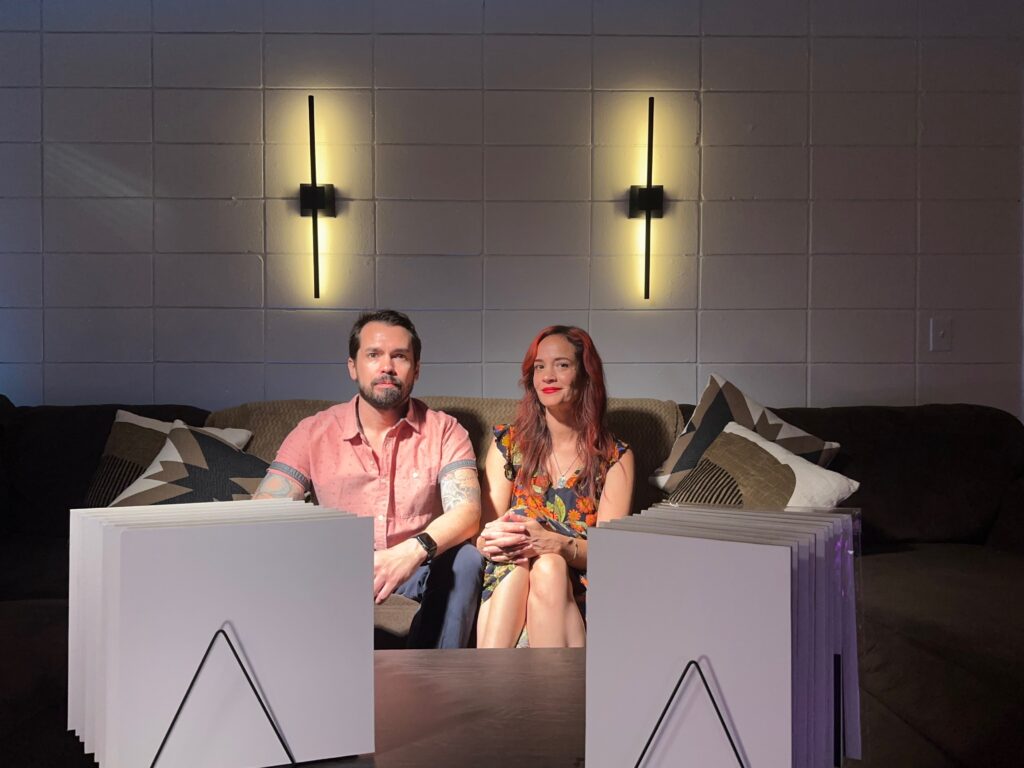 Eric and Jolene seated on a studio sofa behind stacks of white, birch-mounted art panels, illustrating why we started making our own art panels.