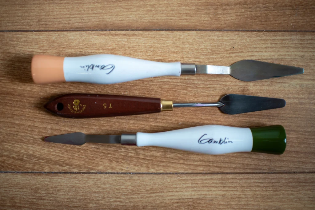 Best Oil Painting Supplies: My Must-Have Tools for Brushes, Mediums, Surfaces & More 2025 2 Three professional palette knives with wood and ergonomic handles laid out on wood grain background