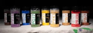 Nine clear jars of powdered artist pigments with black lids lined up on a white cloth against a dark background, each labeled by pigment code (e.g. PBr7, PV16, PB29, PG17, PY37, PY43, PBr7, PR108, PBr7) showing colors from brown and violet through blue, green, yellow, ochre, and red.